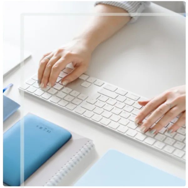 A pair of hands typing on a white keyboard, resting on a clean, white surface. Nearby are a notebook and other blue accessories, suggesting a modern workspace.