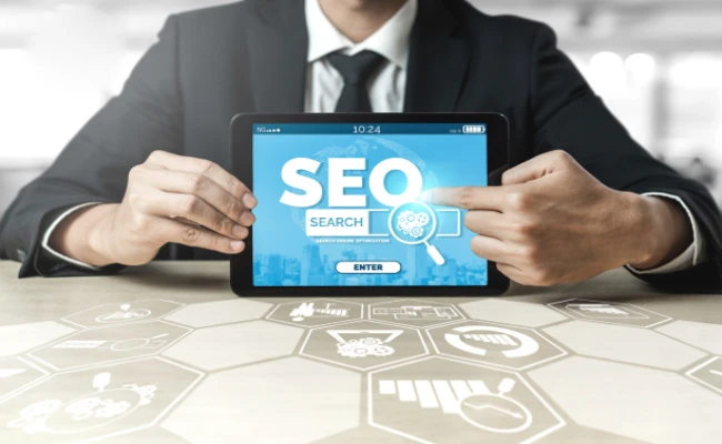 A businessperson holding a tablet displays SEO software. The screen reads “SEO”, “Search” and “Enter”, with a magnifying glass icon. The table has additional SEO icons on it
