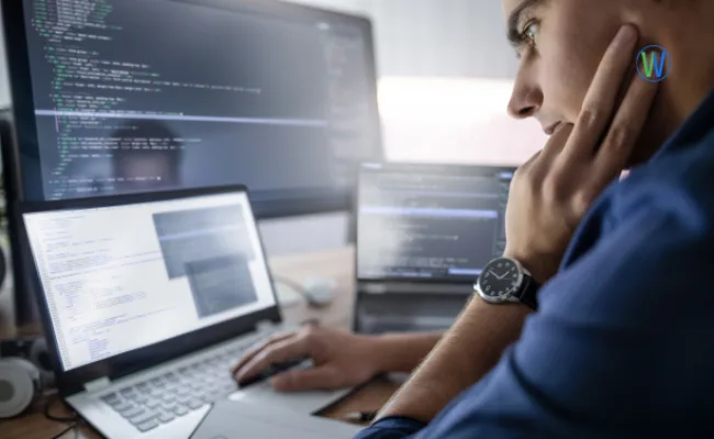 A focused programmer works on multiple screens, possibly coding, with a hand thoughtfully placed near his face. His watch suggests he is on a tight schedule.
