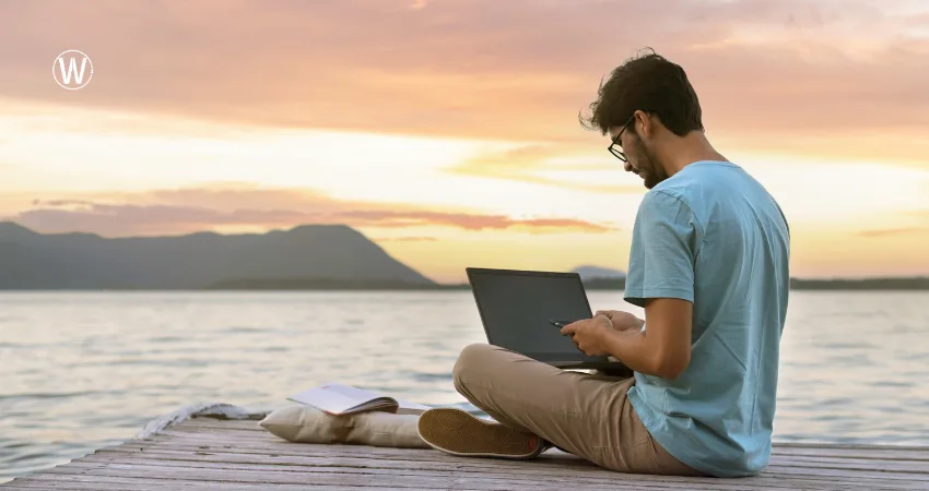 يجلس شخص على رصيف خشبي، يعمل على حاسوبه المحمول. خلفه، تغرب الشمس فوق الماء، مُصدرةً وهجًا برتقاليًا دافئًا.