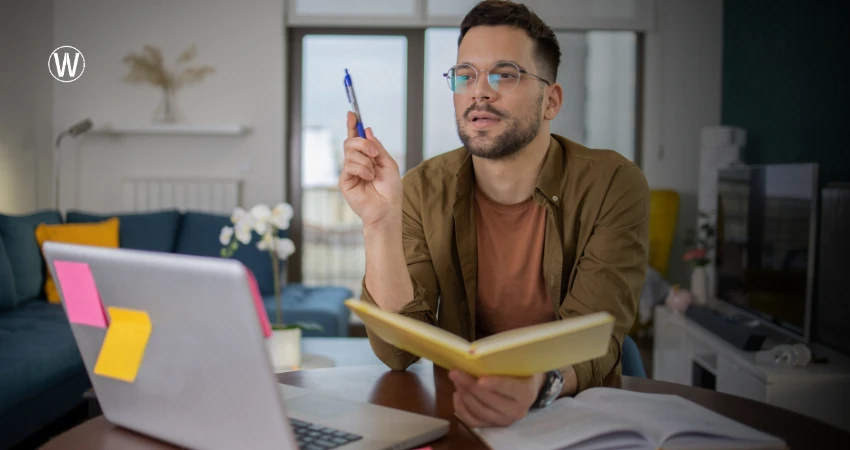 رجل يرتدي نظارات يجلس على مكتب مع حاسوب محمول ودفتر ملاحظات، رافعًا قلمًا كأنه يُثبت وجهة نظر. خلفه، غرفة معيشة بأريكة وديكورات.