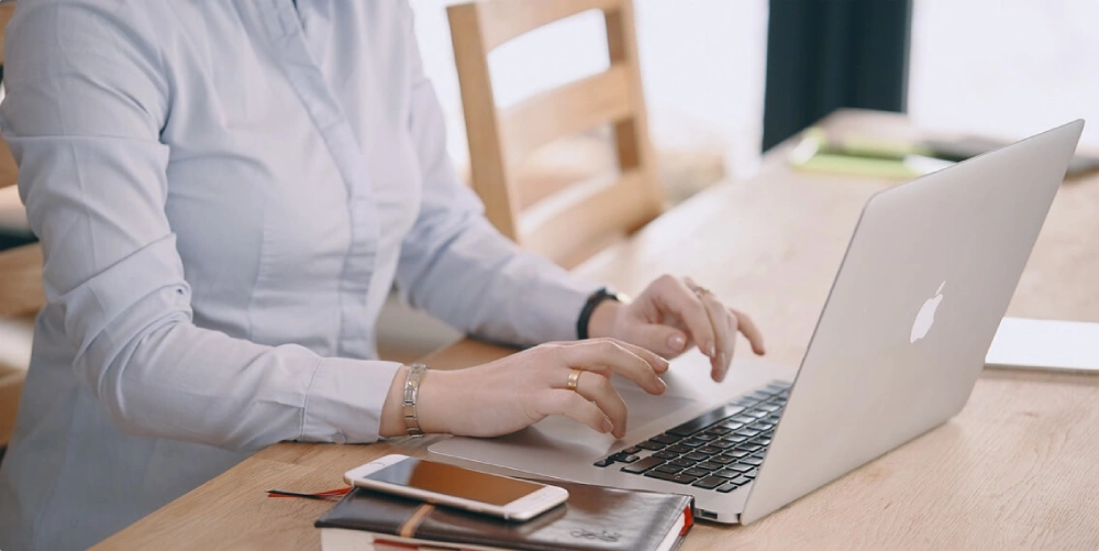 شخص يكتب على حاسوب محمول، وبجانبه هاتف ودفتر ملاحظات على طاولة خشبية. يرتدي قميصًا أزرق فاتحًا بأكمام طويلة، ويضع سوارًا فضيًا على معصمه.