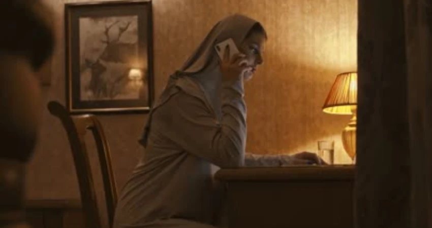 في غرفة ذات إضاءة خافتة، تظهر امرأة محجبة تتحدث عبر الهاتف. تجلس على مكتب قرب مصباح وصورة مؤطرة.