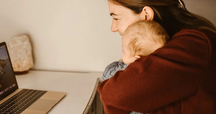 أمٌّ تجلس على مكتبٍ وطفلها بين ذراعيها، تنظر إلى شاشة حاسوبها المحمول. المشهد دافئٌ وجذاب، وبجانب الحاسوب المحمول مصباحٌ ملحيّ.