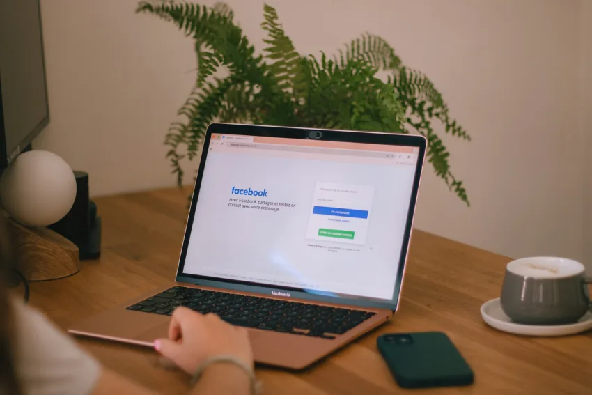 شخص يعمل على جهاز ماك بوك إير، وتظهر صفحة تسجيل الدخول إلى فيسبوك على الشاشة. بجانب الحاسوب، يوجد فنجان قهوة وهاتف. تقول صفحة تسجيل الدخول إلى فيسبوك: "مع فيسبوك، شارك وأبقِ على اتصال مع أصدقائك".