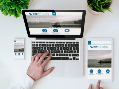 جهاز كمبيوتر محمول موضوع على مكتب بجوار هاتف محمول وجهاز لوحي، يعرض تصميم الويب. الأيدي ظاهرة على لوحة المفاتيح والجهاز اللوحي. النص الظاهر على الشاشات هو "تصميم الويب".