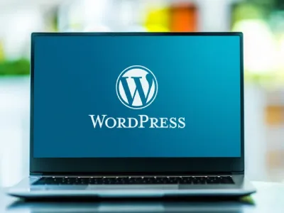 جهاز كمبيوتر محمول مفتوح في المنتصف، يُظهر شاشة زرقاء. تظهر كلمة "WordPress" بخط أبيض كبير أسفل شعار WordPress، وهو حرف "W" أبيض داخل دائرة بيضاء. الكمبيوتر المحمول موضوع على سطح أملس، يعكس الضوء. يظهر خلف الكمبيوتر المحمول محيط غير واضح، بألوان خضراء وصفراء، مما يشير إلى بيئة خارجية. الإضاءة طبيعية، تسلط الضوء على الشاشة وتبرز الشعار. التصوير قريب، يركز على الشاشة، مع تركيز أقل على السطح المحيط. النمط حديث، بلمسة تقنية. الصورة تعطي شعورًا بالوضوح والتركيز على برنامج WordPress.