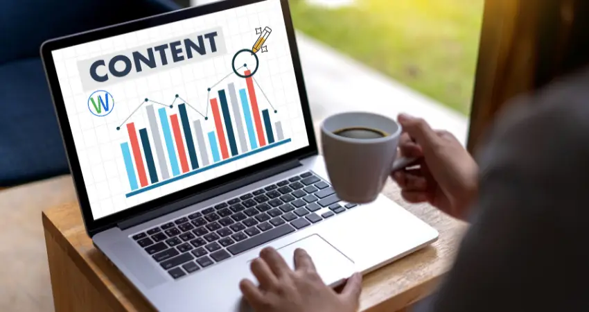 في الصورة، يظهر لابتوب مفتوح على طاولة خشبية. شاشة اللابتوب تعرض كلمة "CONTENT" بخط كبير وواضح، بالإضافة إلى رسم بياني يوضح زيادة في شكل أعمدة ملونة وخطوط متصلة، مع رمز WordPress في الزاوية اليسرى السفلية. شخص، غير محدد الجنس، يظهر نصفه العلوي من الخلف، يمسك بفنجان قهوة في يده اليمنى، ويداه على لوحة مفاتيح اللابتوب. يبدو أن الشخص يتابع الرسم البياني. الإضاءة طبيعية وتأتي من نافذة على اليمين. الإطار العام ضيق ويبرز تركيزًا على موضوع العمل والتحليل. الألوان دافئة، مع هيمنة الألوان الزرقاء والبرتقالية في الرسم البياني. الجو يوحي بالتركيز والعمل، وربما تحليل المحتوى أو البيانات.