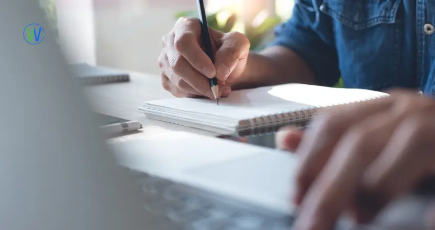 رجل يكتب في دفتر ملاحظات موضوع على طاولة. تظهر يداه، مع مسكة يد يمنى قلم رصاص أسود، وهو يكتب على صفحة بيضاء فارغة. يرتدي قميصًا أزرقًا. الدفتر ذو حلقات حلزونية. يقع الدفتر في منتصف الصورة، بينما تظهر يد أخرى و جزء من حاسوب محمول في الخلفية. الإضاءة طبيعية، مع تركيز على الأيدي و الدفتر. يظهر ضوء خفيف قادم من أعلى الصورة، مما يبرز تفاصيل القلم و الدفتر. تظهر الصورة بأسلوب واقعي و تعبر عن التركيز و العمل.