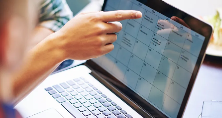 يشير شخص إلى تقويم على شاشة حاسوب محمول. يعرض التقويم الاجتماعات والتقارير.