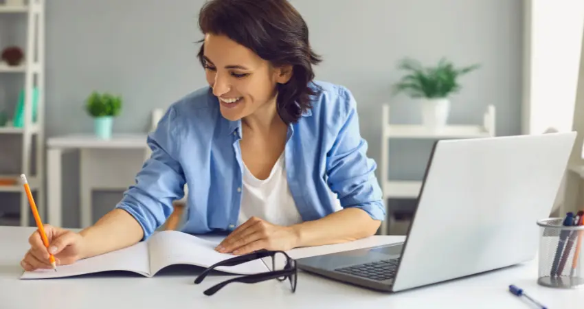 امرأة شابة ذات بشرة فاتحة وشعر أسود قصير، في منتصف العمر، تجلس أمام مكتب أبيض. ترتدي قميصاً أزرق فاتح مفتوحاً على قميص داخلي أبيض. تعبر عن السعادة والتركيز، وتبتسم وتنظر إلى الأسفل نحو دفتر مفتوح تكتب فيه بقلم رصاص. على الطاولة أمامها، توجد نظارات سوداء مفتوحة، وجهاز كمبيوتر محمول رمادي اللون. يظهر خلف الكمبيوتر كوب أقلام. على يمينها، توجد نبتة خضراء صغيرة في وعاء. الخلفية عبارة عن جدار رمادي فاتح مع نباتات أخرى وقطعة أثاث بيضاء في الخلف. الإضاءة طبيعية، تخلق جواً هادئاً ودافئاً. يتم التقاط الصورة من زاوية منخفضة بعض الشيء، مع التركيز على المرأة وعملها.