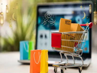 عربة تسوق صغيرة محملة بالطرود بالقرب من جهاز كمبيوتر محمول. تظهر حقائب ملونة وزينة تحمل طابع رمضان، بالإضافة إلى عبارة "رمضان كريم".