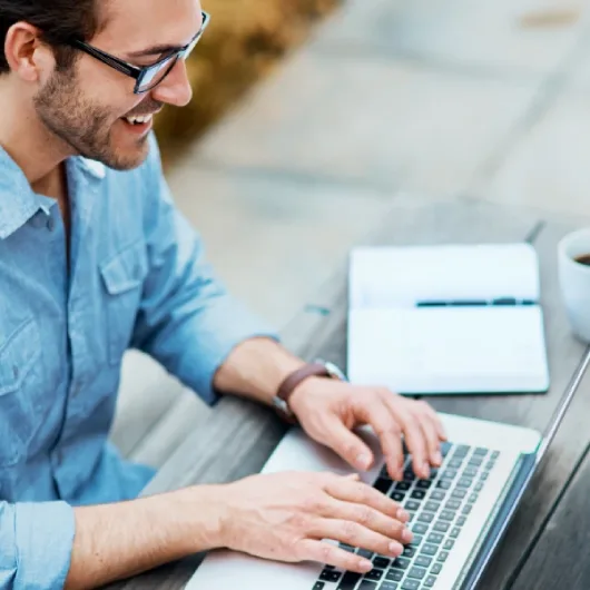 رجل يبتسم وهو يقوم بالتدوين على حاسوبه المحمول على طاولة خارجية، حاملاً دفتر ملاحظات وكوبًا. يرتدي الرجل نظارات وقميصًا أزرق فاتحًا بأزرار.