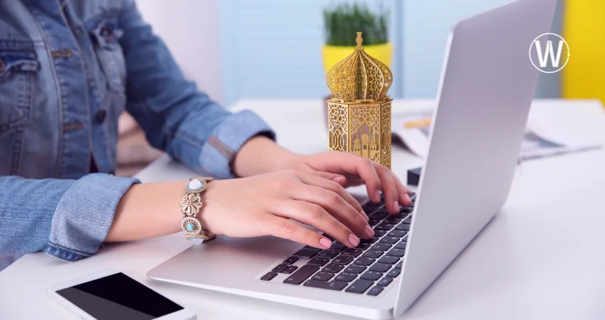 امرأة ترتدي سترة جينز تكتب على جهاز كمبيوتر محمول. يوجد هاتف ذكي على الطاولة البيضاء بالقرب من الكمبيوتر المحمول، وخلفه فانوس رمضان ذهبي مزخرف.