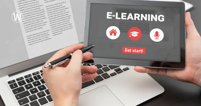 صورة مقربة لشخصين، أحدهما يمسك جهازًا لوحيًا والآخر بقلم، فوق جهاز كمبيوتر محمول. يظهر على الجهاز اللوحي كلمة "E-LEARNING" بخط أبيض عريض، وتحتها رموز حمراء: منزل، قبعة تخرج، ميكروفون، مع زر "ابدأ!" أحمر. اليد اليمنى تمسك بالجهاز اللوحي