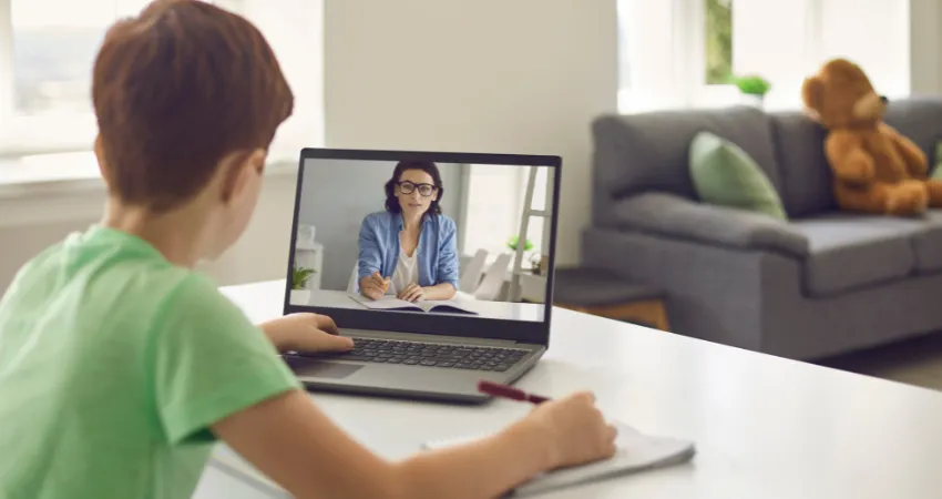طفل يحضر مؤتمراً عبر الفيديو مع معلمه باستخدام جهاز كمبيوتر محمول، بينما يدون ملاحظات في دفتر ملاحظات بقلم. في الخلفية تظهر أريكة ودب صغير.