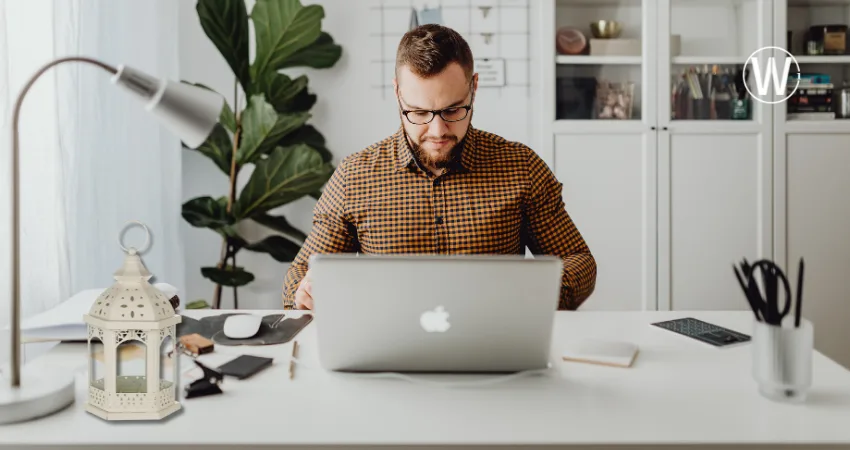 يجلس رجل على مكتب يعمل على جهاز كمبيوتر محمول. المكتب مغطى بمصباح وفانوس رمضان مزخرف ولوازم مكتبية. توجد نبتة خلفه.