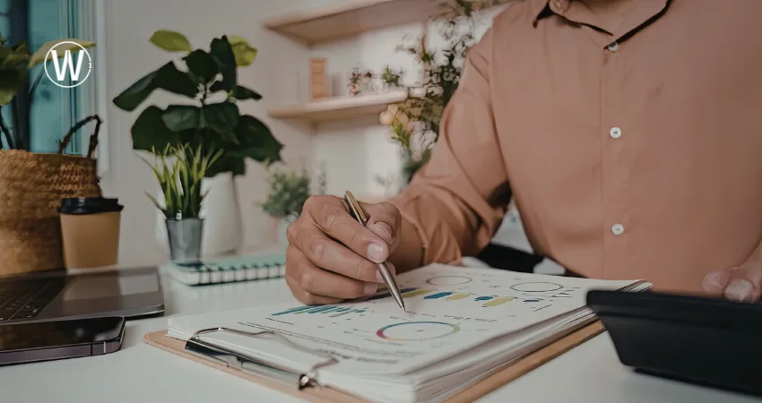 يقوم شخص بمراجعة مخطط عملي لرمضان تحتوي على رسوم بيانية, مستخدماً قلماً وآلة حاسبة. ويحتوي المكتب على جهاز كمبيوتر محمول ونباتات ومشروب ساخن.