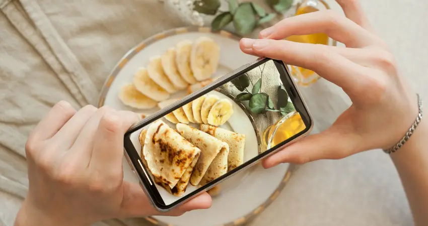 تمسك يدٌ بهاتف لالتقاط صورة لطبق إفطار. يحتوي الطبق على فطائر وموز مقطع.