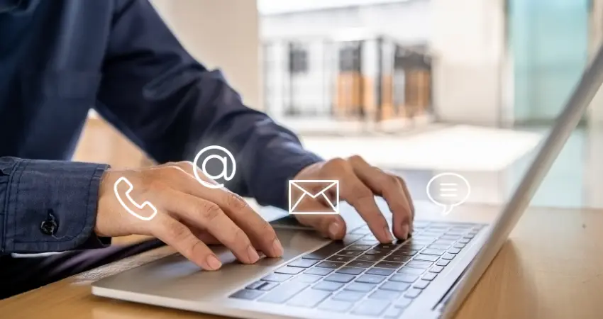 شخص يكتب على جهاز كمبيوتر محمول، مع وجود أيقونات الهاتف والبريد الإلكتروني والرسائل فوق يديه وجهاز الكمبيوتر. تشير الصورة إلى طرق متعددة للتواصل مع شخص ما.