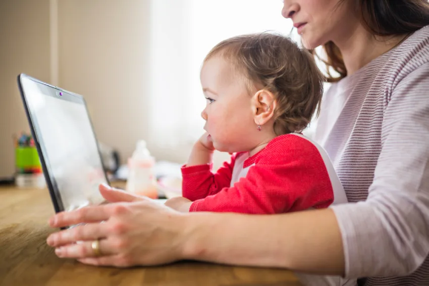 طفل صغير بعمر حوالي سنة يجلس على حضن امرأة، كلاهما ينظران إلى شاشة حاسوب محمول. يرتدي الطفل قميصًا أحمر وأبيض، ولديه شعر أشقر مجعد ووجه مستدير، مع تعبير هادئ. تبدو بشرة الطفل بيضاء، مع قرط في الأذن. المرأة، تظهر جزئيا، ترتدي قميصًا مقلمًا باللونين الأبيض والرمادي، وتبدو بشرتها بيضاء أيضًا. يقع الطفل في الجزء الأوسط من الصورة، متجهاً إلى اليسار، بينما تظهر المرأة من الكتفين إلى الأعلى على اليمين. الحاسوب المحمول يقع على طاولة خشبية. خلفهما، هناك زجاجة رضاعة، مما يوحي ببيئة منزلية. الإضاءة طبيعية، مع التركيز على الوجهين، مما يخلق جواً دافئاً وحميمياً. الخلفية غير واضحة، مع ألوان محايدة. الصورة ذات منظور قريب، مما يركز على العلاقة بين الطفل والمرأة.