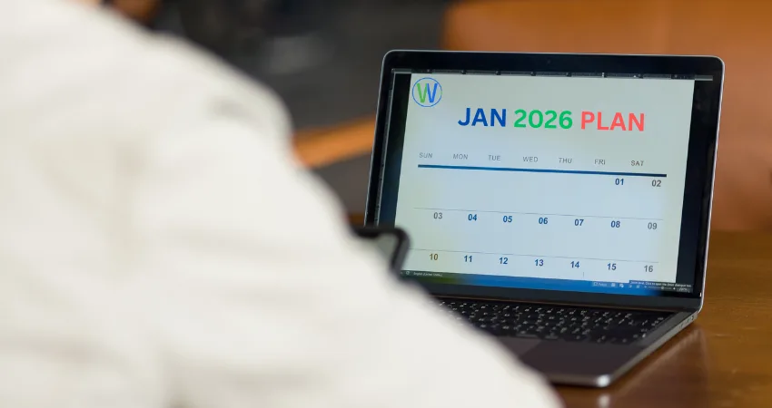شخص يرتدي سترة بيضاء في المقدمة، ظهرت فقط كتفيه ورأسه في غير بؤرة التركيز، يمسك بهاتف أسود. في مركز الصورة، يوجد جهاز كمبيوتر محمول مفتوح على طاولة خشبية. شاشة الكمبيوتر تعرض خطة شهر يناير 2026، مكتوبة بألوان الأزرق والأخضر والأحمر. يظهر شعار (W) الأزرق في الزاوية العلوية اليسرى للشاشة. يظهر تقويم بسيط مع أيام الأسبوع والأرقام. الإضاءة طبيعية، مع ظلال طفيفة. المنظور من وجهة نظر الشخص الذي يمسك بالهاتف، ويوحي بأنه يراقب التقويم. الجو العام عملي ومهني.