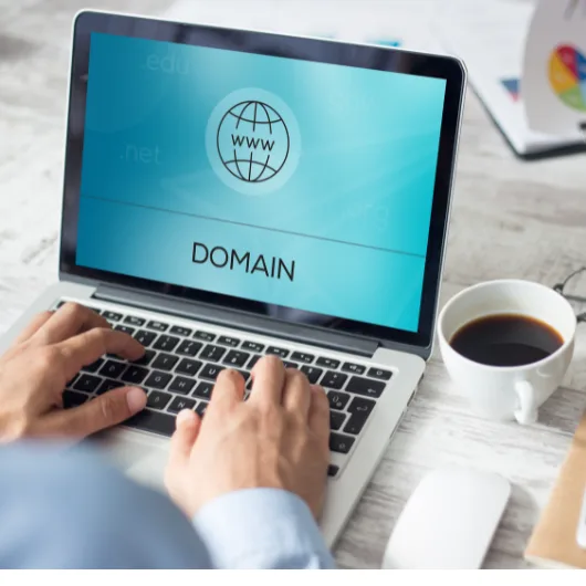 تطبع يداها على جهاز كمبيوتر محمول يعرض كلمة "اسم النطاق" باللون الأزرق، مع وجود كرة أرضية وكلمة "www" و".com" و".net. "في المنتصف. ويوجد كوب قهوة وفأرة على المكتب بجانب الكمبيوتر.