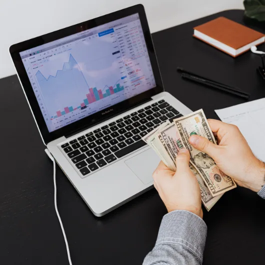 شخص يحمل نقوداً أمام جهاز كمبيوتر محمول، وعلى الشاشة يظهر رسم بياني للأسهم,توضح ان اختيار النيتش المربح اساس الربح من التدوين والمشهد عبارة عن مكتب عمل مع ملحقات إضافية