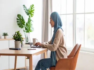 امرأة ترتدي حجاباً أزرق تمارس العمل من المنزل على مكتب خشبي فاتح اللون. يحتوي المكتب على جهاز كمبيوتر محمول، وكوب، وهاتف، ونباتات منزلية موضوعة بجوار نافذة مشرقة.