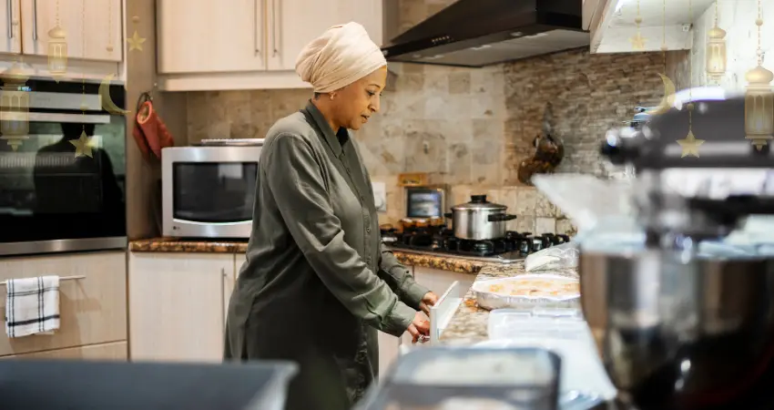 امرأة ترتدي الحجاب تقف في المطبخ. تقف عند طاولة عليها أجهزة منزلية، وهي تعد وجبات افطار رمضان