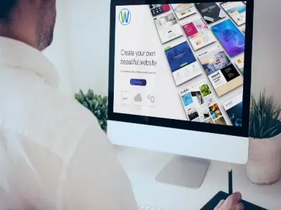 رجل في منتصف العمر يجلس أمام شاشة كمبيوتر. يظهر وجهه جزئياً، مع رؤية من الخلف. يرتدي قميصاً أبيضاً. الشاشة تعرض واجهة لموقع ويب، مع نص "Create your own beautiful website" وشعار W باللونين الأبيض والأزرق. تعرض الشاشة أيضاً العديد من تصاميم صفحات الويب. توجد نبتة صغيرة في إناء على يمين الشاشة. يمسك الرجل بقلم على لوحة الرسم الرقمية، والتي تقع على الطاولة أمامه. الإضاءة طبيعية، تخلق ظلالًا ناعمة. البيئة مكتبية حديثة مع تصميم نظيف وأبيض. تتركز الصورة على الشاشة مع التركيز على تصميم الويب المعروض.