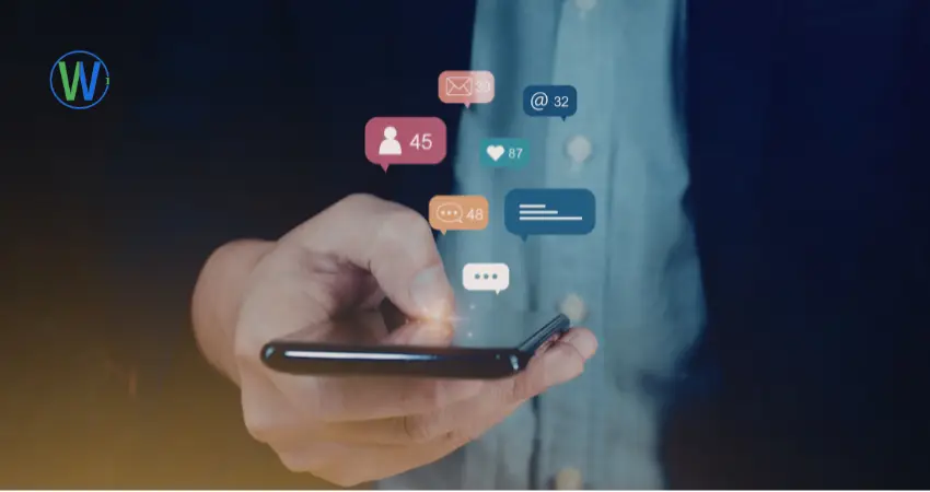 يد تمسك بهاتف ذكي أسود، مع ظهور أيقونات وسائل التواصل الاجتماعي فوق الهاتف. الشخص في الصورة يرتدي قميصاً أزرق فاتح. الخلفية داكنة مع بقع زرقاء وذهبية. يظهر الضوء بشكل أساسي على الهاتف واليد. تظهر أيقونات وسائل التواصل الاجتماعي في شكل فقاعات الكلام الملونة، بما في ذلك أيقونة ملف شخصي مع رقم "45"، أيقونة رسالة بريد إلكتروني مع "52"، أيقونة قلب مع "87"، أيقونة الدردشة مع "48"، وأيقونة تعليق مع ثلاثة نقاط وأيقونة "@32". وجهة النظر مقربة، مع تركيز حاد على اليد والهاتف. الإضاءة مركزة، مما يخلق تبايناً مع الخلفية الداكنة. الجو العام حديث ومرتبط بالتكنولوجيا ووسائل التواصل الاجتماعي. شعار "W" في الدائرة الزرقاء الخضراء موجود في الزاوية اليسرى العليا.